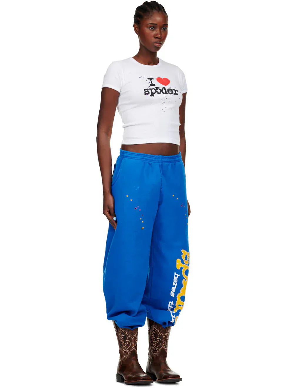 A woman stands against a plain background wearing a white crop top, Sp5der Sweatpants – Unisex Blue Cotton Sweatpants with Yellow Side Logo and Star Print, and brown cowboy boots.