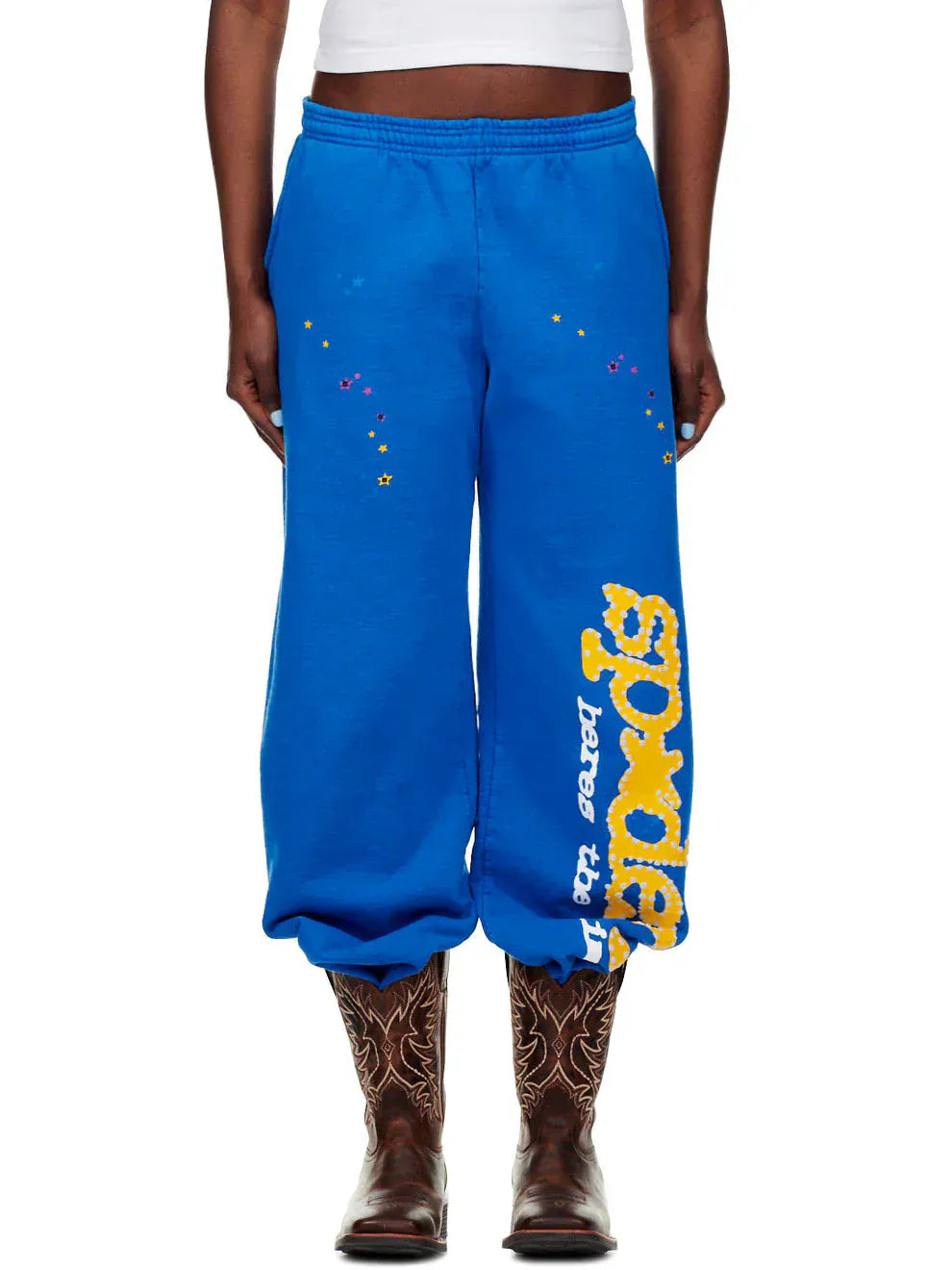 Styled for a bold look, these Sp5der Sweatpants – Unisex Blue Cotton Sweatpants with Yellow Side Logo and Star Print are paired with brown cowboy boots and a cropped white top, perfect for standout streetwear jogger style.