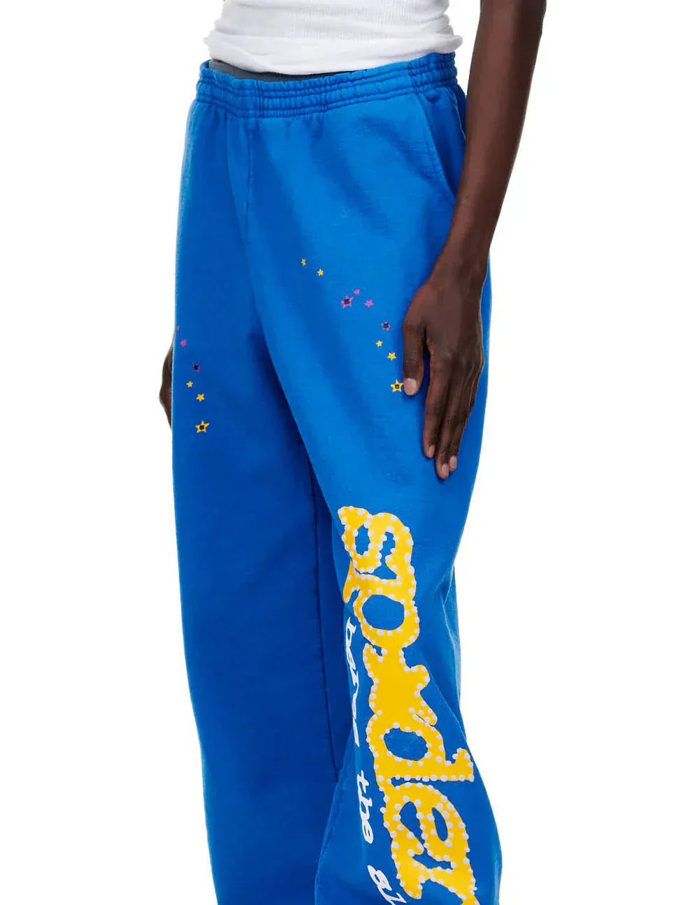 A person in Sp5der Sweatpants – Unisex Blue Cotton Sweatpants with Yellow Side Logo and Star Print, posing casually; ideal cotton streetwear joggers for any style.