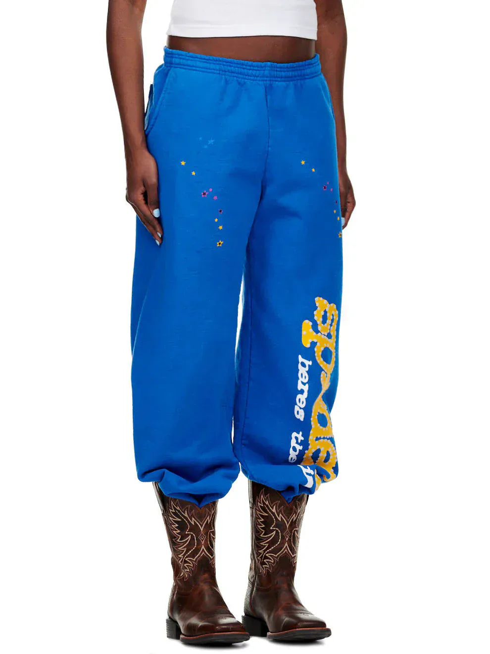 A person in Sp5der Unisex Blue Cotton Sweatpants with a yellow side logo and star print, a white crop top, and brown cowboy boots stands against a white background.