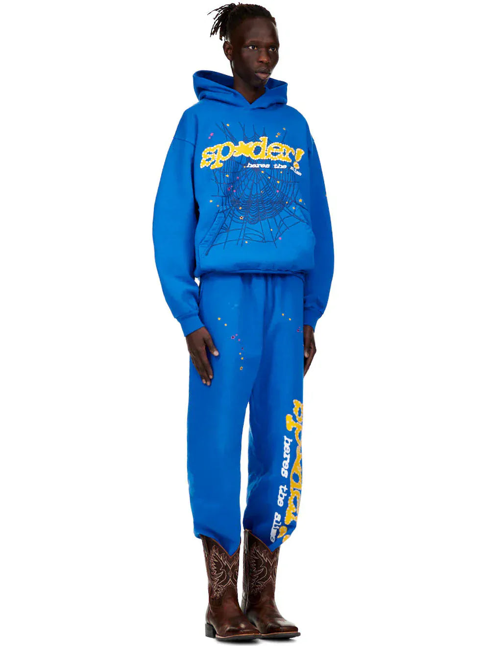 A person wearing a blue graphic hoodie and Sp5der Sweatpants – Unisex Blue Cotton Sweatpants with Yellow Side Logo and Star Print, paired with brown cowboy boots.