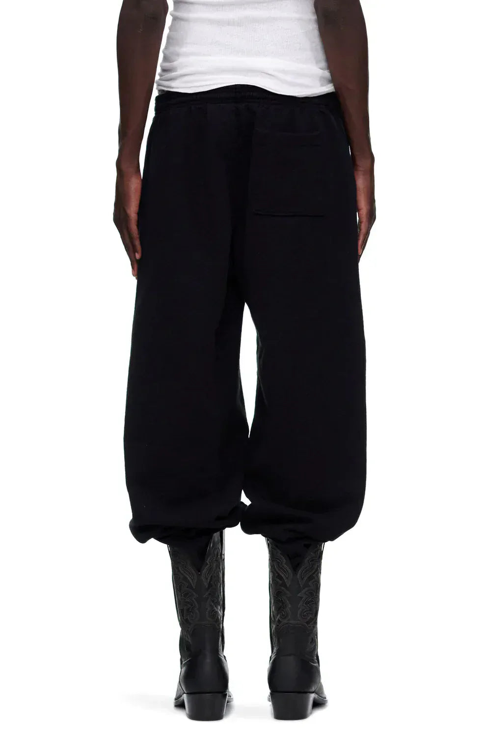 Person wearing Sp5der Sweatpants—unisex black cotton sweatpants with pink graphic logo and star print—paired with black boots and a white tank top, shown from the back.