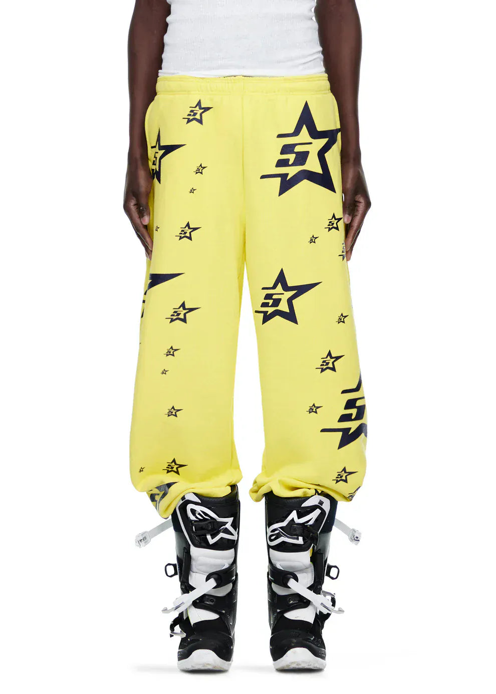 Person wearing Sp5der Sweatpants—unisex yellow cotton with all-over star print—paired with a white ribbed shirt and black motocross boots for a standout streetwear look.
