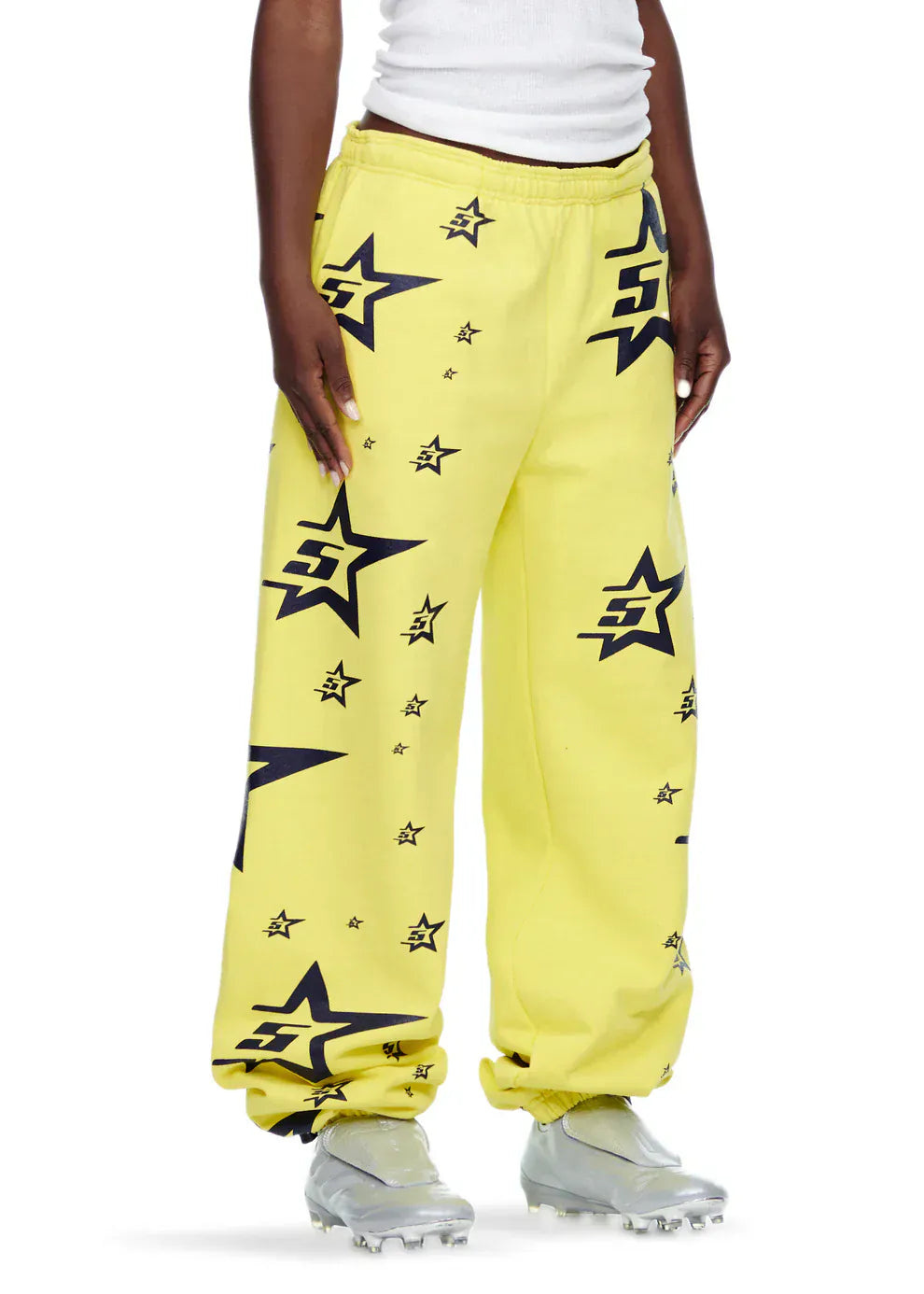 A person wears Sp5der Sweatpants – Unisex Yellow Cotton Sweatpants with All-Over Star Print, paired with a white tank top and silver athletic cleats for a standout streetwear look.