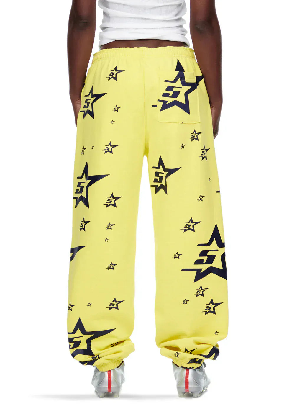A person is seen from the back wearing Sp5der Sweatpants – Unisex Yellow Cotton Sweatpants with an all-over star print, featuring an elastic waistband, paired with white sneakers and a white top for a streetwear look.