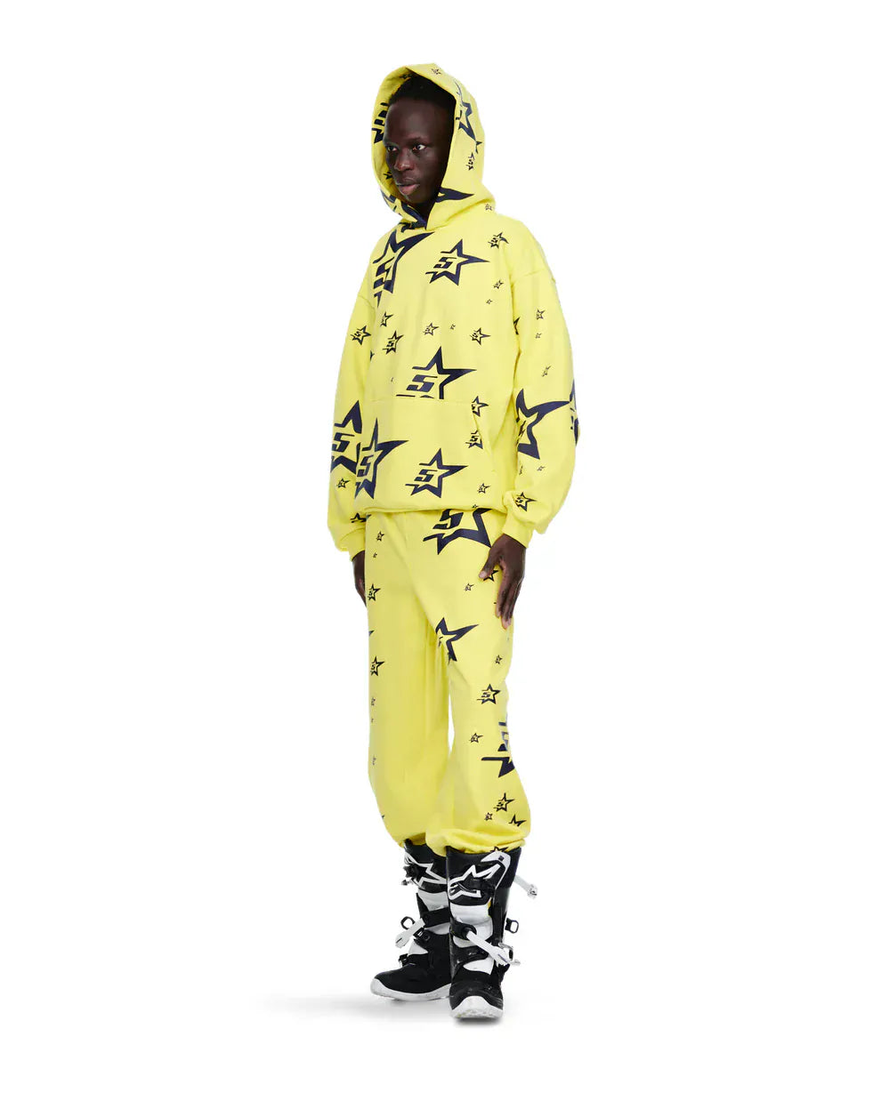 Wearing a yellow hoodie and Sp5der Sweatpants – Unisex Yellow Cotton Sweatpants with All-Over Star Print – paired with black and white boots, this person stands against a white background, exemplifying bold streetwear style.