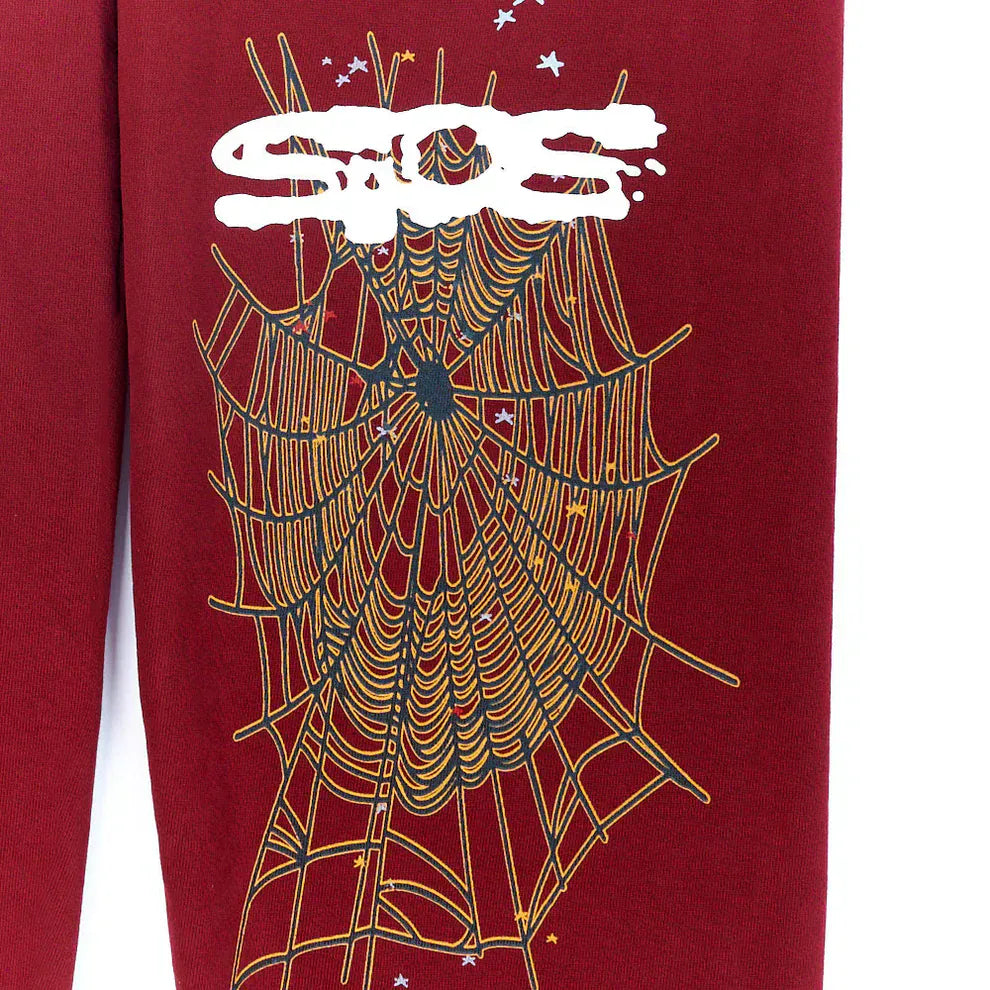 The Sp5der Sweatpants are unisex burgundy cotton sweatpants featuring a large gold spider web graphic, small white stars, and bold graffiti-style text—ideal for standout streetwear looks.