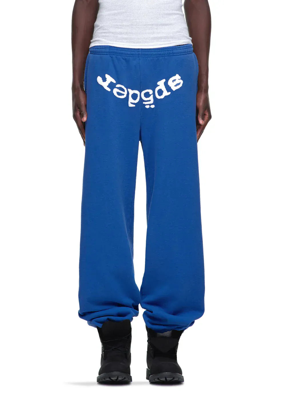 Person wearing unisex blue Sp5der Sweatpants with a bold white front logo print, paired with a white tank top and black shoes—a standout streetwear look.