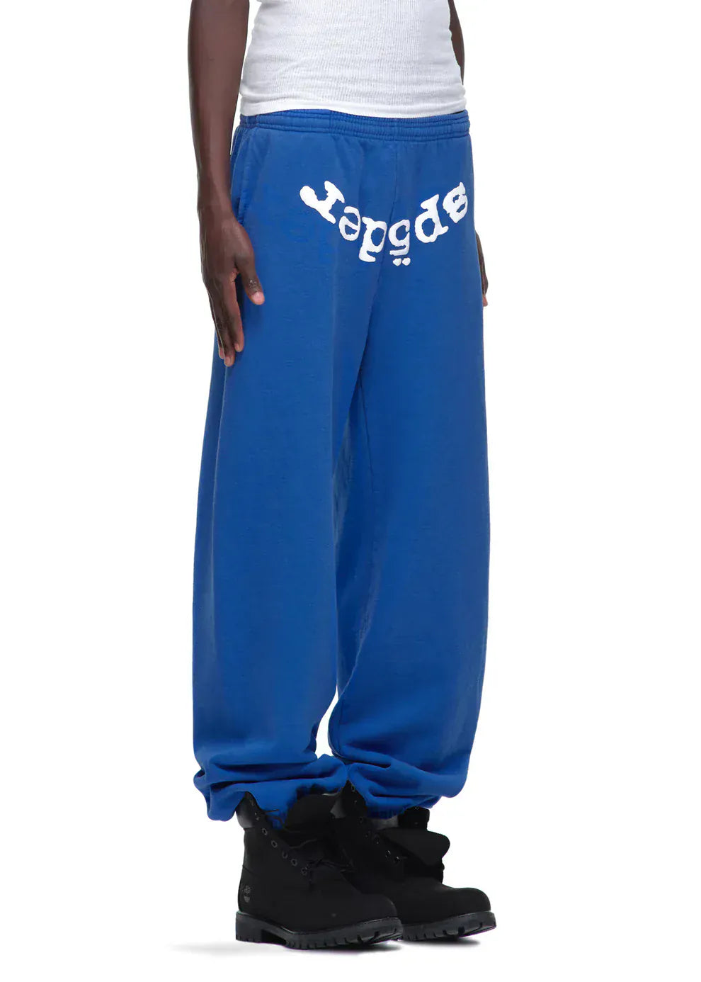 Sporting Sp5der Sweatpants – unisex blue cotton pants with a front logo print – along with a white tank top and black boots, this person stands against a plain background, embodying effortless streetwear style.