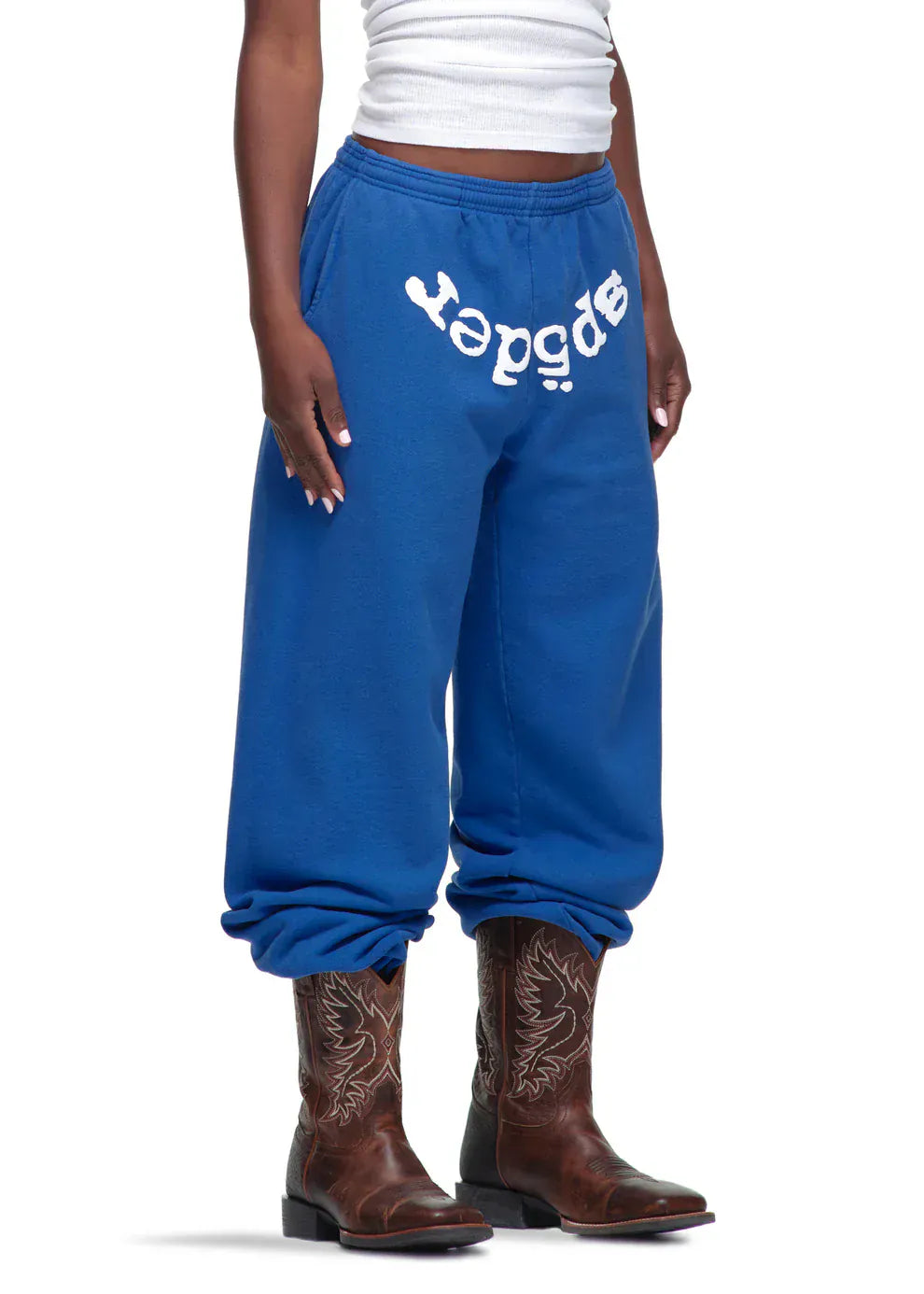 Wearing Sp5der Sweatpants – Unisex Blue Cotton Sweatpants with a front logo print, a white tank top, and brown cowboy boots, someone stands against a plain background, merging streetwear and western styles.