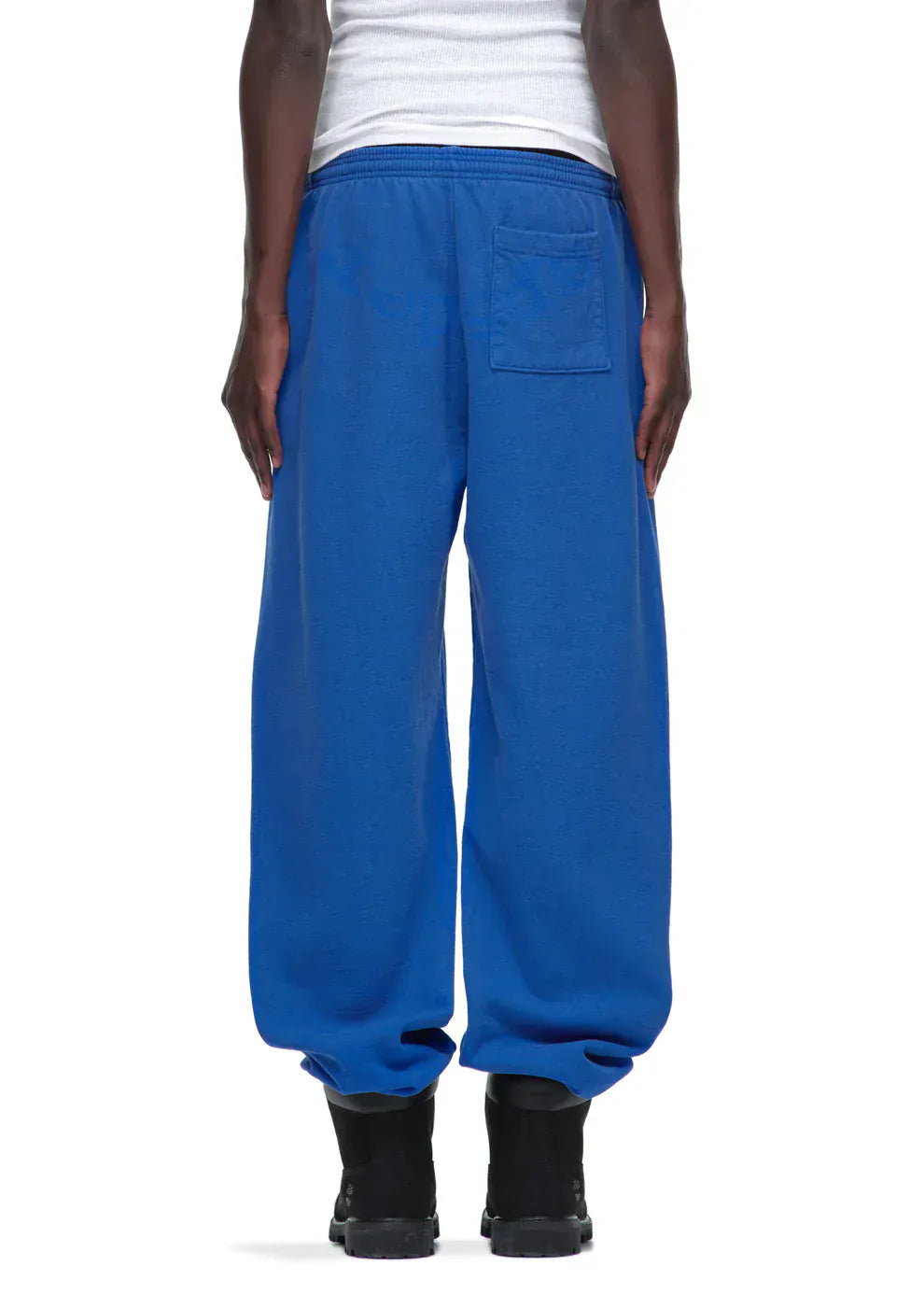 A person is shown from the back in Sp5der Sweatpants – unisex blue cotton sweatpants with a front logo print, paired with black shoes and a white sleeveless top, capturing a relaxed streetwear vibe.