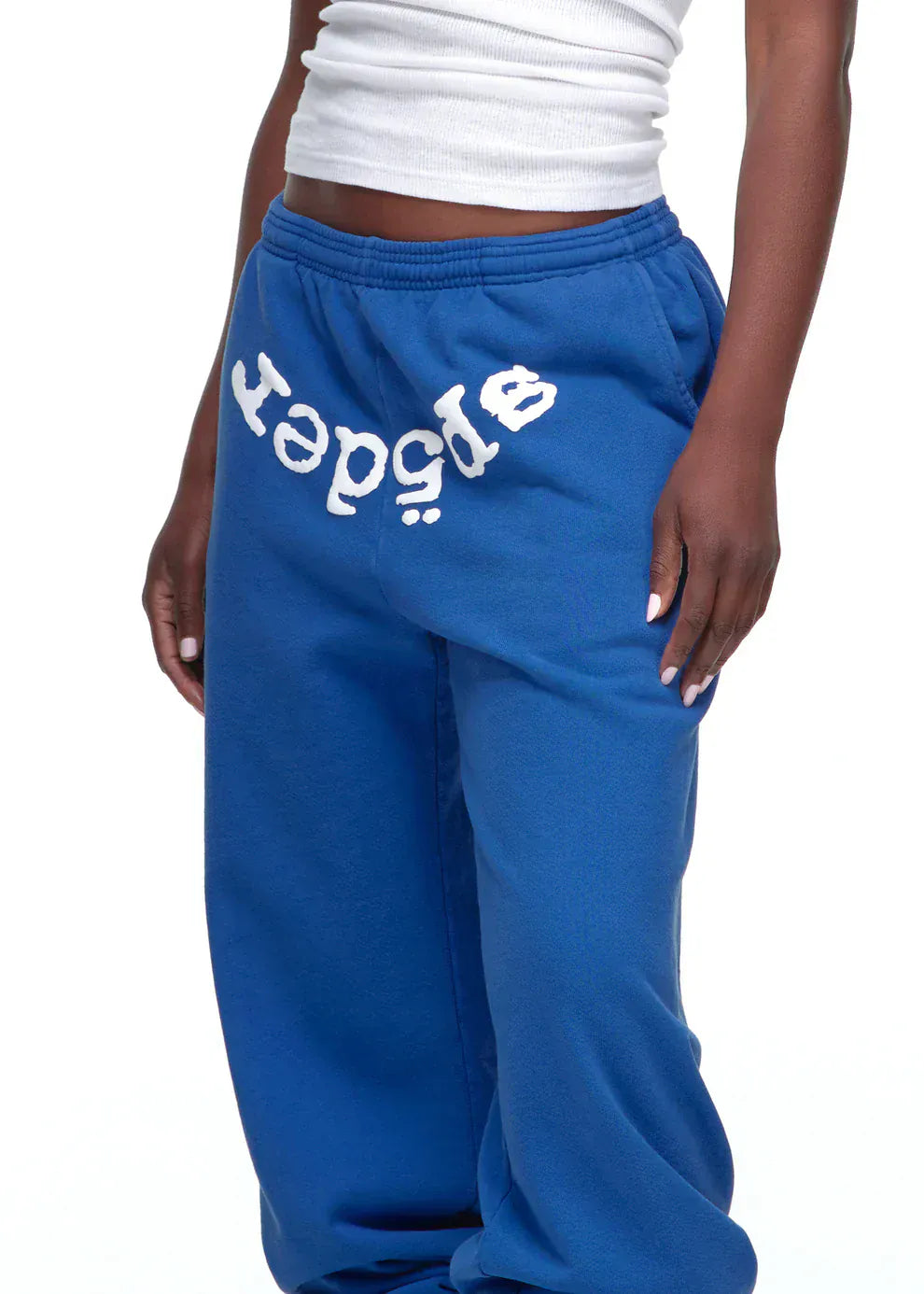 A person in blue Sp5der Sweatpants – Unisex Blue Cotton Sweatpants with a front logo print, paired with a white ribbed tank top for a casual streetwear look.