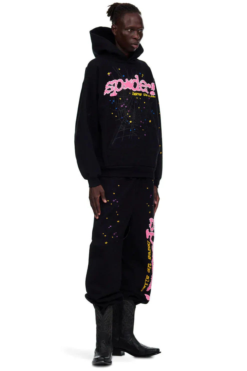 A person wears Sp5der Sweatpants—unisex black cotton pants with a pink graphic logo and star print—paired with a black hoodie and black cowboy boots for a bold streetwear look.
