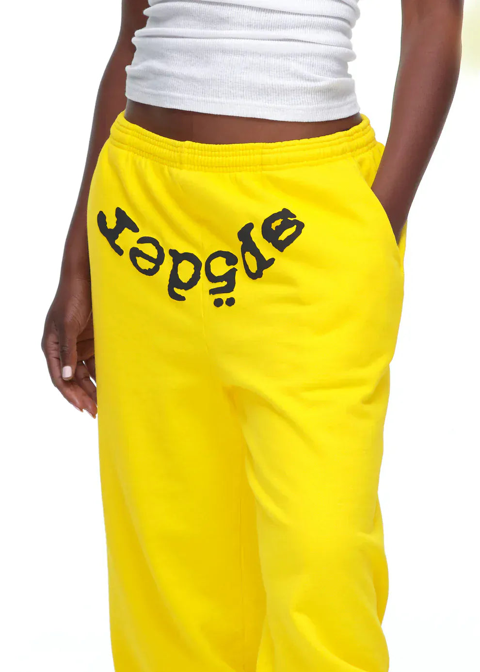Person wearing Sp5der Sweatpants—unisex yellow cotton sweatpants with a bold front logo print—styled with a white ribbed tank top. One hand in the pocket, perfect for streetwear jogger enthusiasts.