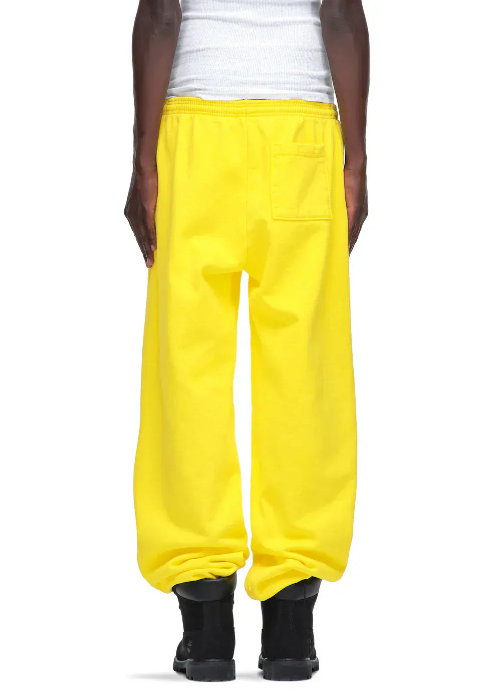 A person stands facing away, wearing a white tank top, black boots, and bold yellow Sp5der Sweatpants – unisex cotton sweatpants with a front logo print.