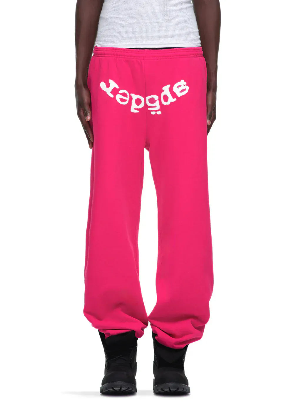 Someone wearing Sp5der Sweatpants – unisex pink cotton sweatpants with a front logo print, paired with black shoes and a white ribbed top.