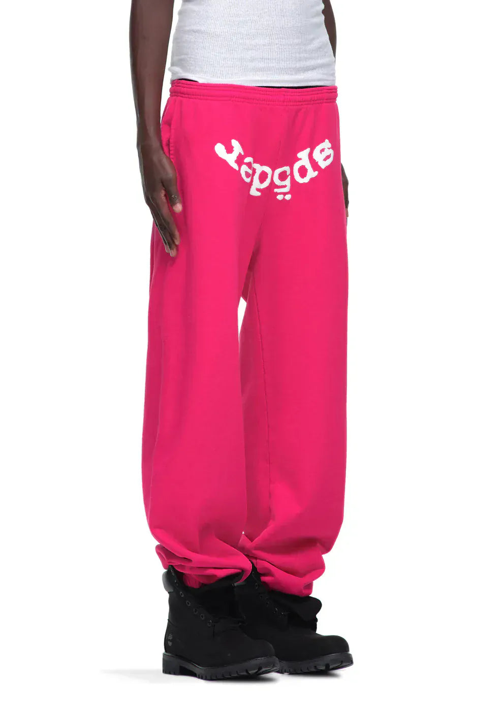 Person wears Sp5der Sweatpants—unisex pink cotton joggers with a front logo print—paired with black boots against a plain background, showcasing bold pink streetwear style.