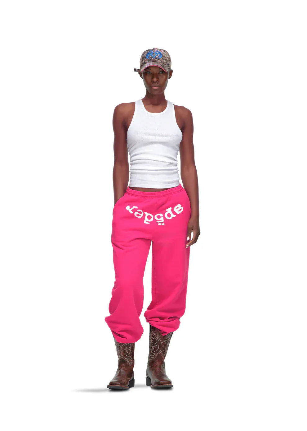 A person wearing a white tank top, Sp5der Sweatpants – Unisex Pink Cotton Sweatpants with Front Logo Print, brown cowboy boots, and a patterned headscarf stands against a white background.