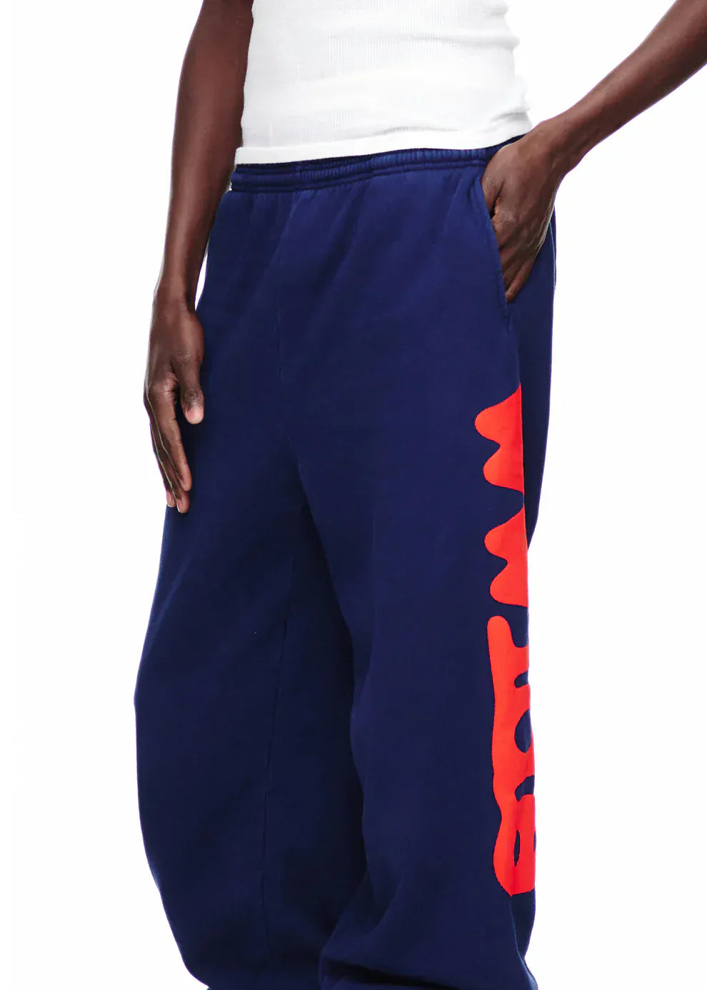 A person in a white tank top and Sp5der Sweatpants Navy Blue Red Side Logo Print—100% cotton streetwear jogger pants—stands with one hand in their pocket.