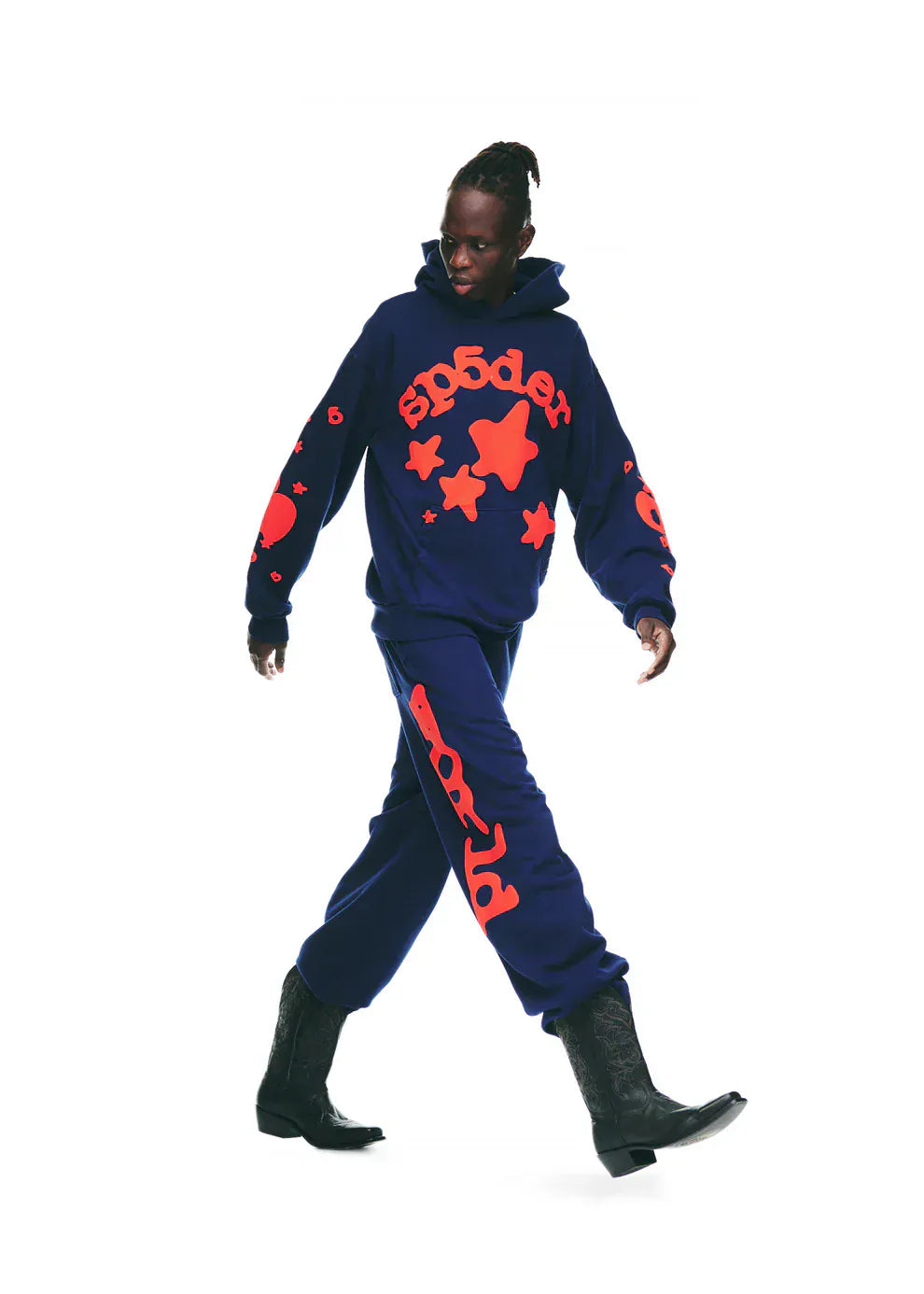 A person wearing Sp5der Sweatpants Navy Blue Red Side Logo Print – 100% Cotton Streetwear Jogger Pants, a navy hoodie with bold red graphics, and black cowboy boots walks against a white background.