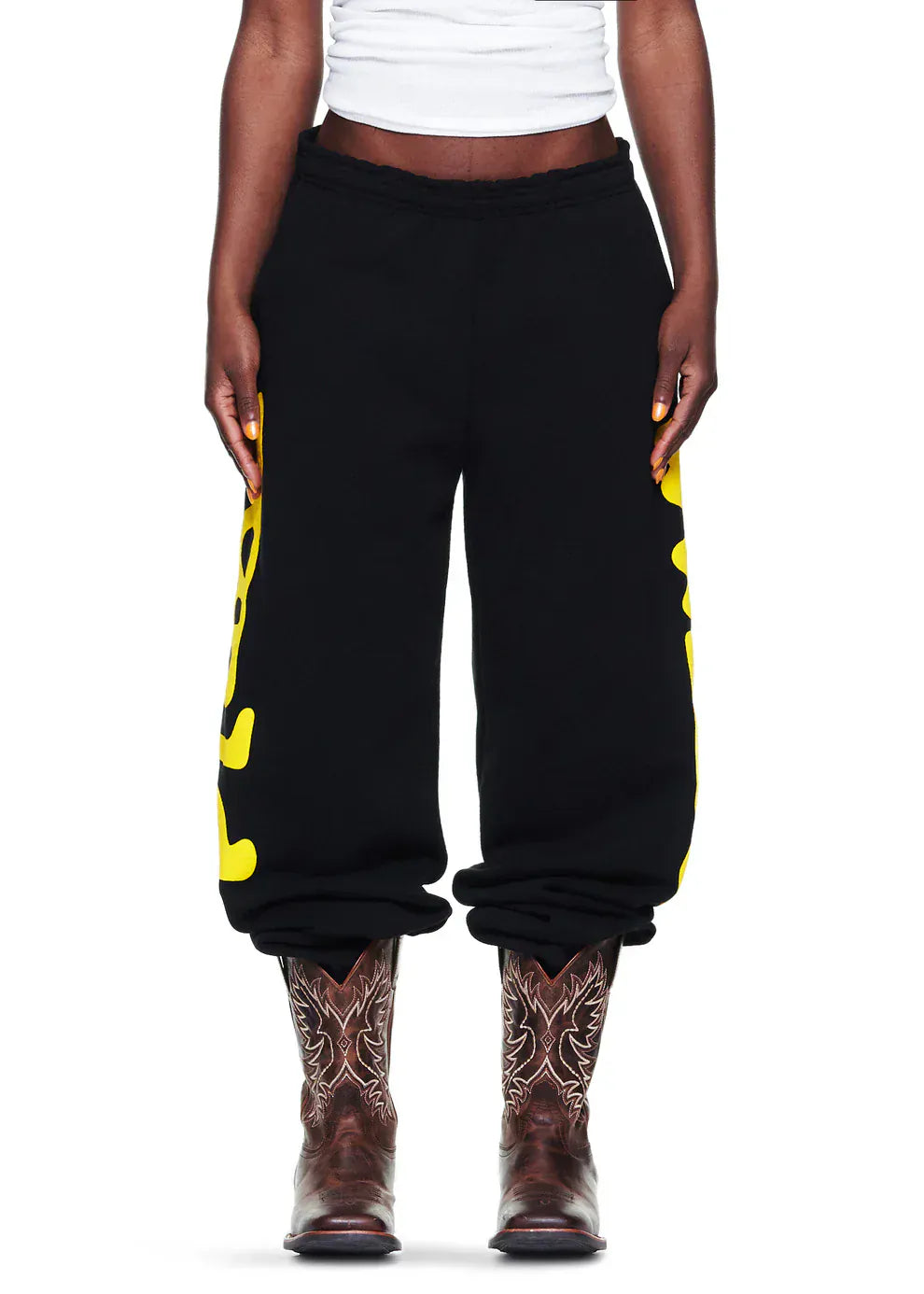 A person wears Sp5der Sweatpants Black Yellow Side Logo Print, a white shirt, and brown cowboy boots—standing against a white background, effortlessly blending streetwear jogger style with classic Western flair.