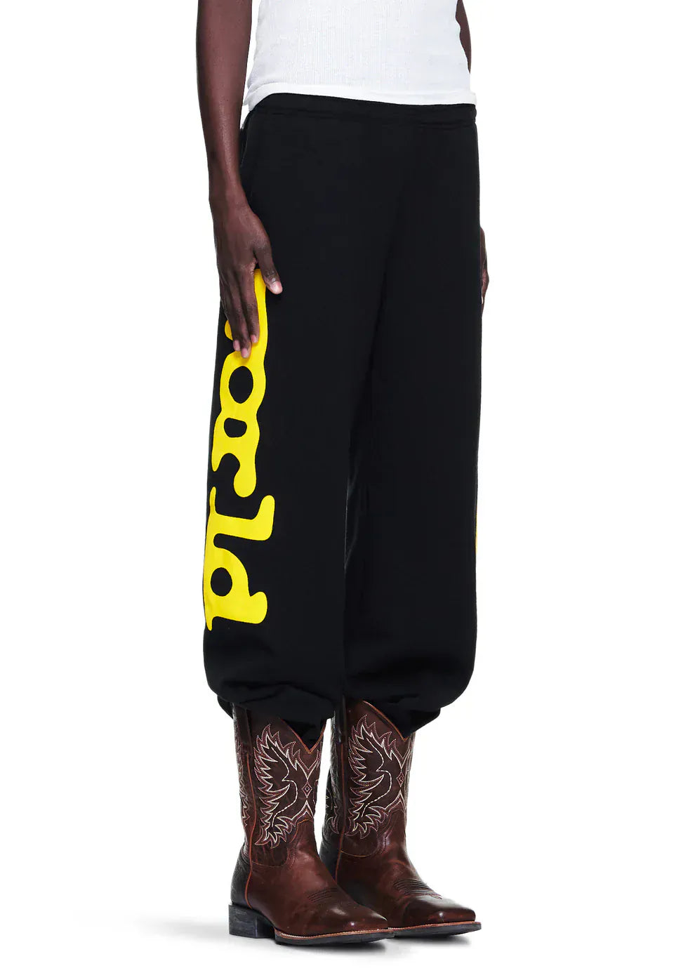 A person wearing Sp5der Sweatpants Black Yellow Side Logo Print – 100% Cotton Streetwear Jogger Pants, paired with brown cowboy boots and a white sleeveless shirt.