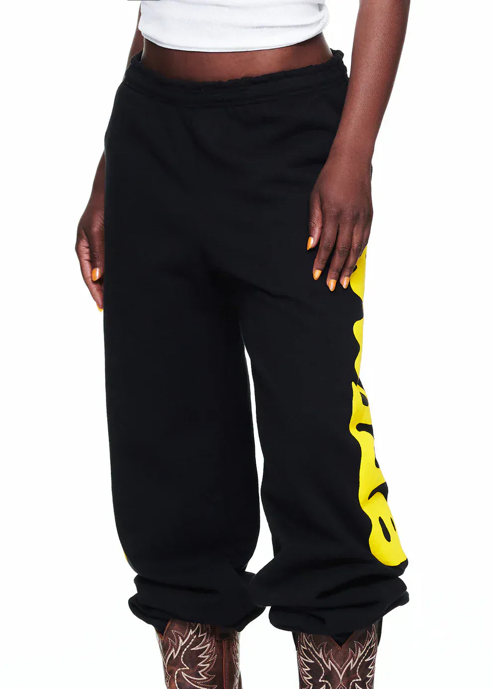 A person wearing Sp5der Sweatpants Black Yellow Side Logo Print – 100% Cotton Streetwear Jogger Pants, paired with a white crop top and patterned tights. Only the torso and legs are visible, highlighting a standout streetwear look.