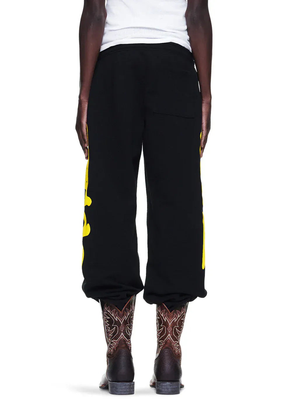 Someone seen from the back wears Sp5der Sweatpants Black Yellow Side Logo Print joggers (100% cotton streetwear), a white shirt, and patterned brown cowboy boots—a striking mix of urban style and Western flair.