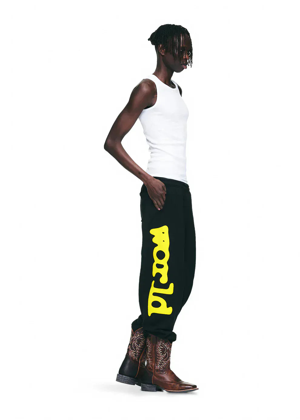 A person wearing a white tank top, Sp5der Sweatpants Black Yellow Side Logo Print – 100% Cotton Streetwear Jogger Pants, and brown cowboy boots stands against a plain white background.