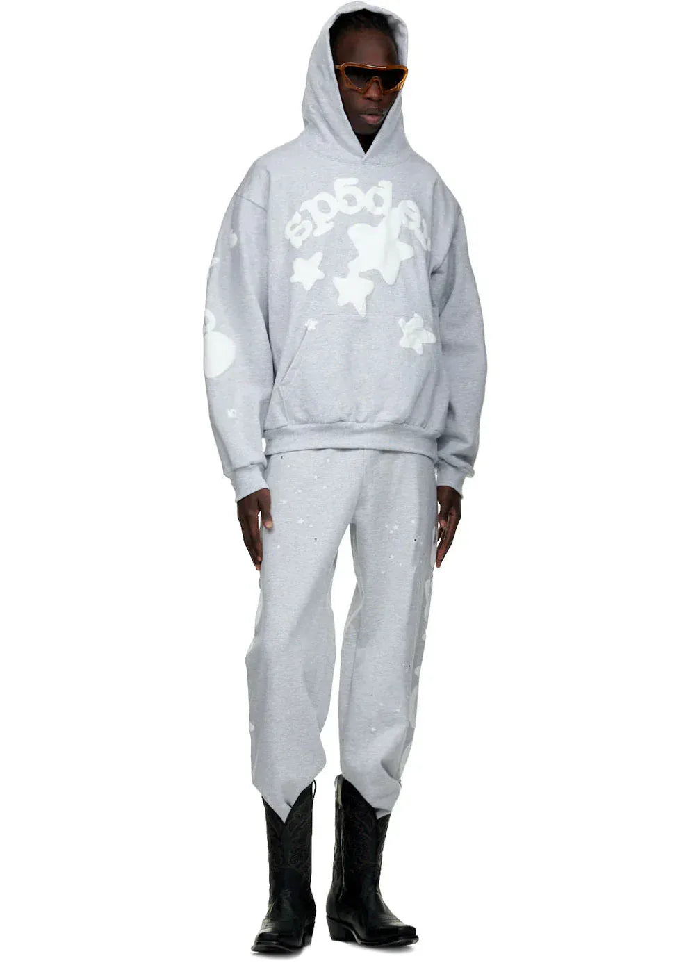 A person wearing a gray hoodie with star designs, 100% Cotton Sp5der Sweatpants for Men – Grey Streetwear Jogger Pants with Graphic Print, black boots, and orange sunglasses stands against a plain background.
