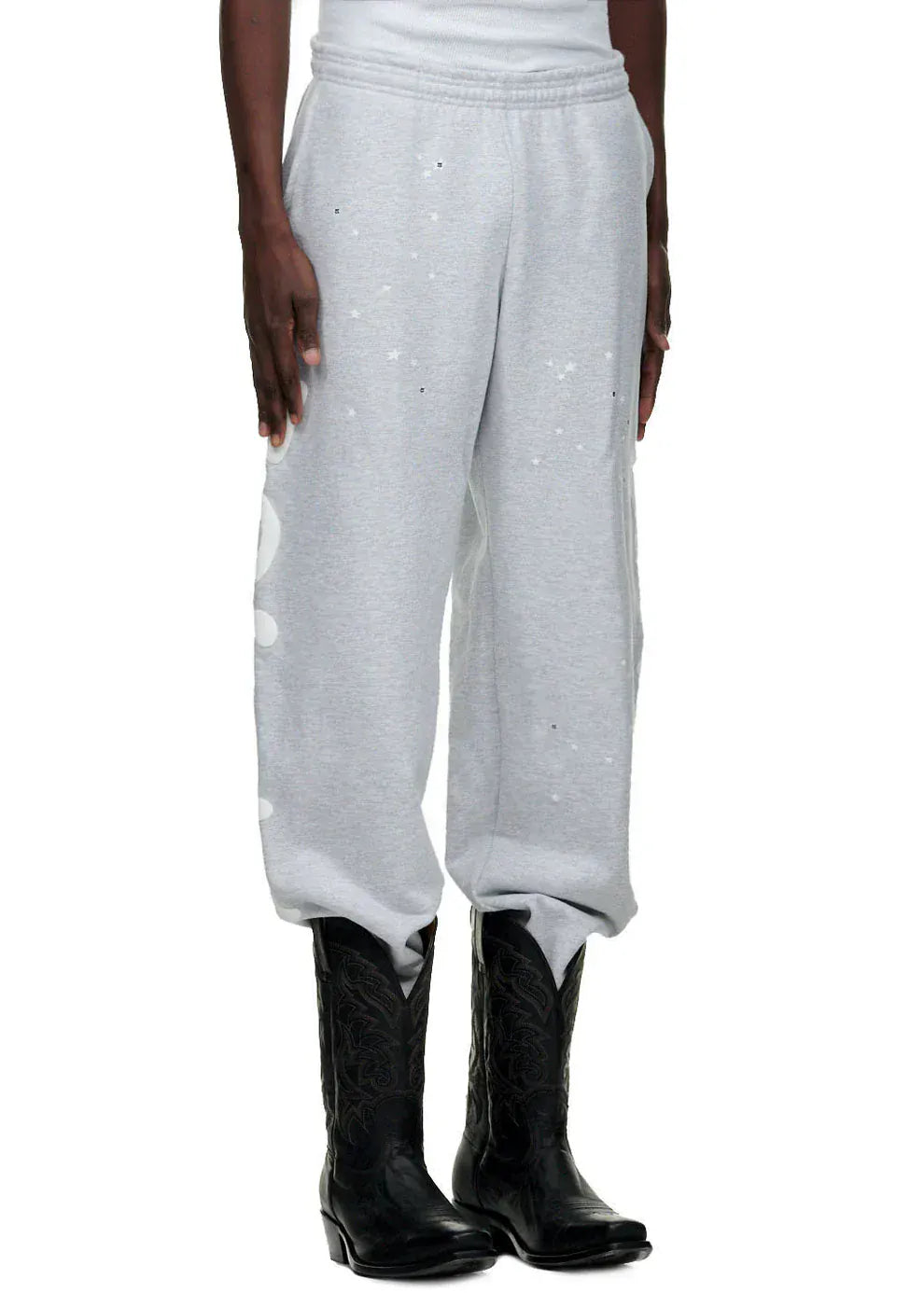 A person wearing 100% Cotton Sp5der Sweatpants for Men – Grey Streetwear Jogger Pants with Graphic Print and black cowboy boots, standing against a white background.