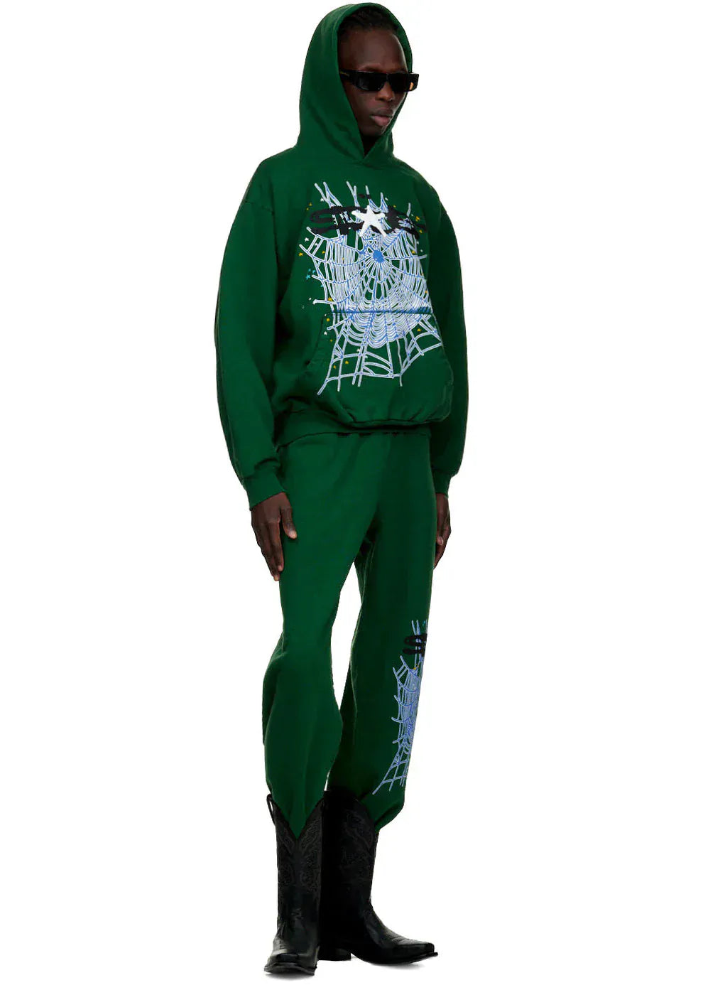 A person in dark green 100% cotton Sp5der Sweatpants for Men with a spider web graphic, styled with a green hoodie, black cowboy boots, and sunglasses against a plain background for bold streetwear jogger vibes.