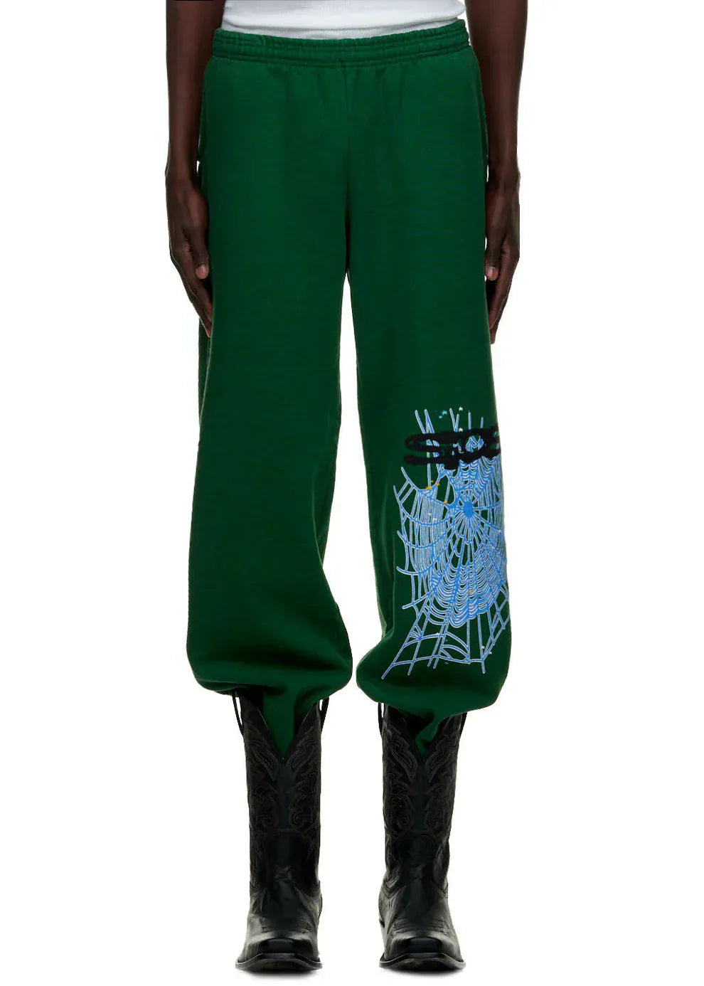 A person wears 100% Cotton Sp5der Sweatpants for Men—dark green streetwear jogger pants with a blue spider web graphic and relaxed fit—paired with black cowboy boots, standing against a white background.
