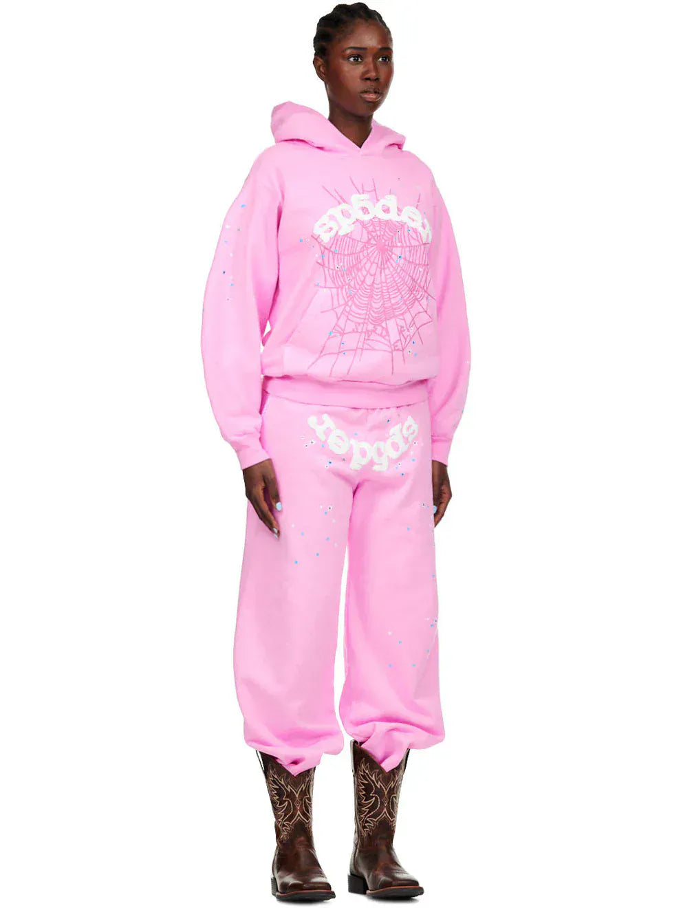 A person in pink streetwear—wearing a pink hoodie and Sp5der Sweatpants (Unisex Light Pink Cotton with Front Logo and Star Print)—paired with brown cowboy boots, stands against a plain background.