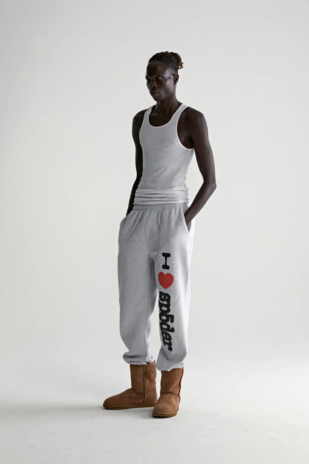 A person stands in a plain studio wearing a gray tank top, 100% Cotton Sp5der Sweatpants for Men—light grey jogger pants with “I Love Sp5der” graphic and relaxed fit—and brown boots, hands in pockets.