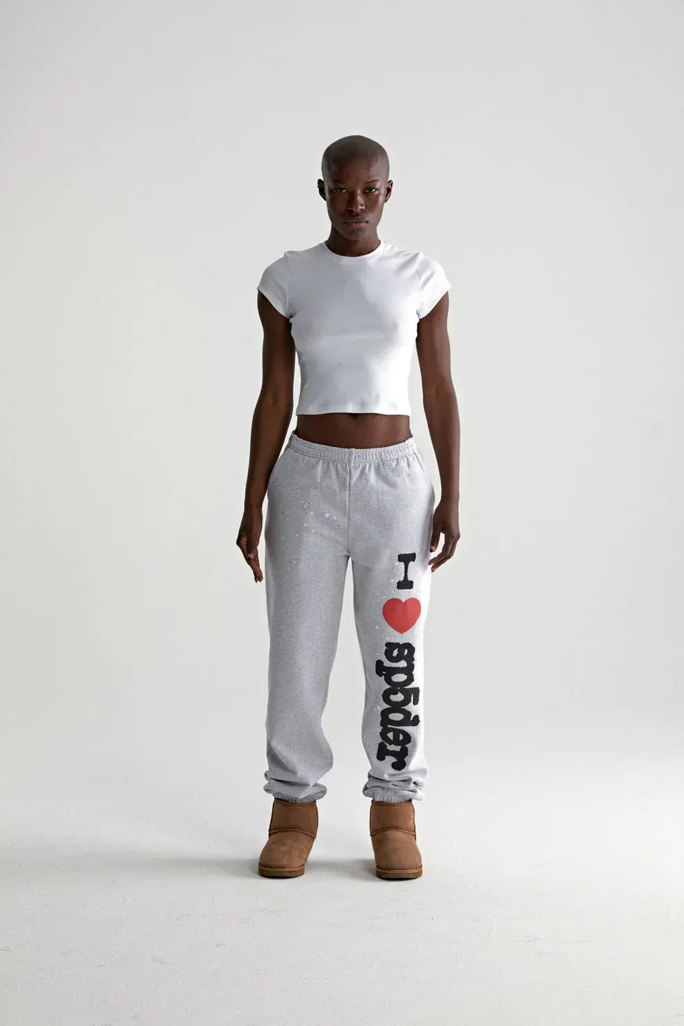 A person stands facing forward in a white cropped t-shirt, light grey 100% cotton Sp5der sweatpants with “I Love Sp5der” graphic, relaxed fit, and brown slippers, against a plain white background.