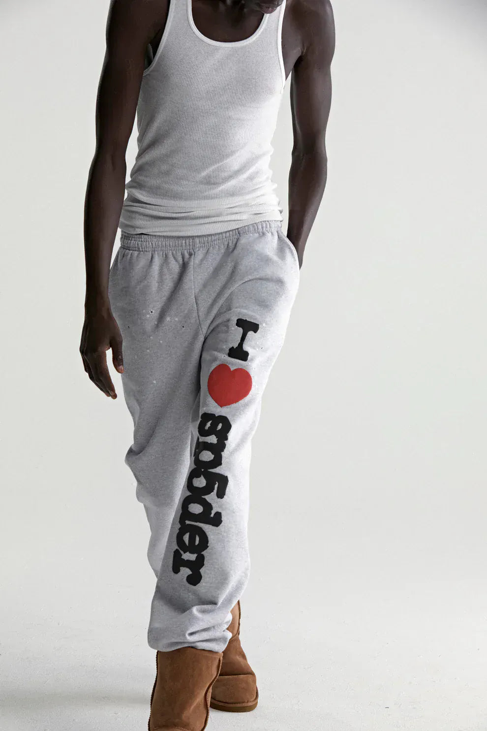 A person in a white tank top, brown boots, and 100% Cotton Sp5der Sweatpants for Men—light grey joggers with “I Love Sp5der” graphic and a relaxed fit—walks against a plain background.