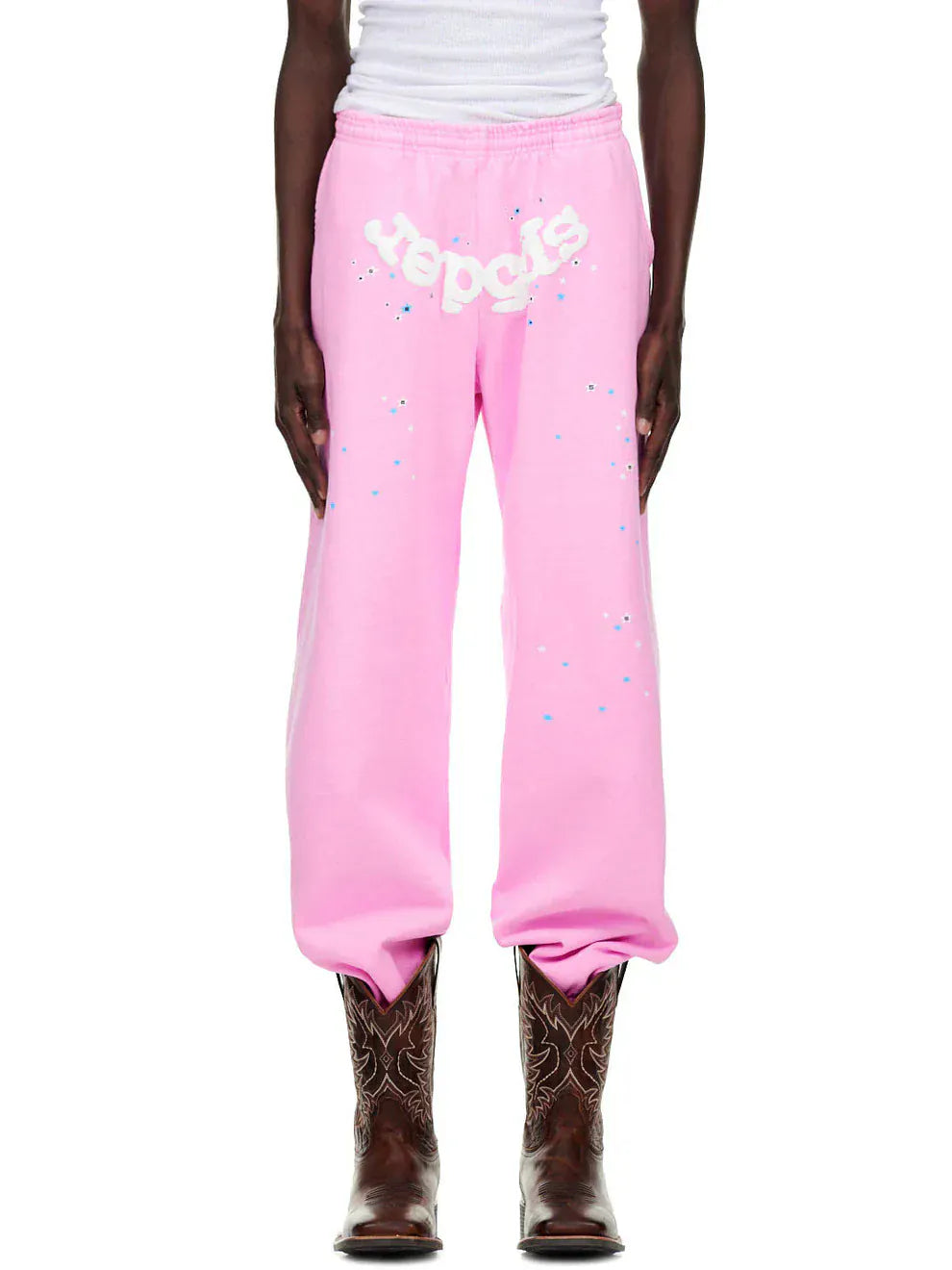 A person wearing Sp5der Sweatpants – Unisex Light Pink Cotton Sweatpants with front logo and star print, a white top, and brown cowboy boots stands against a white background.