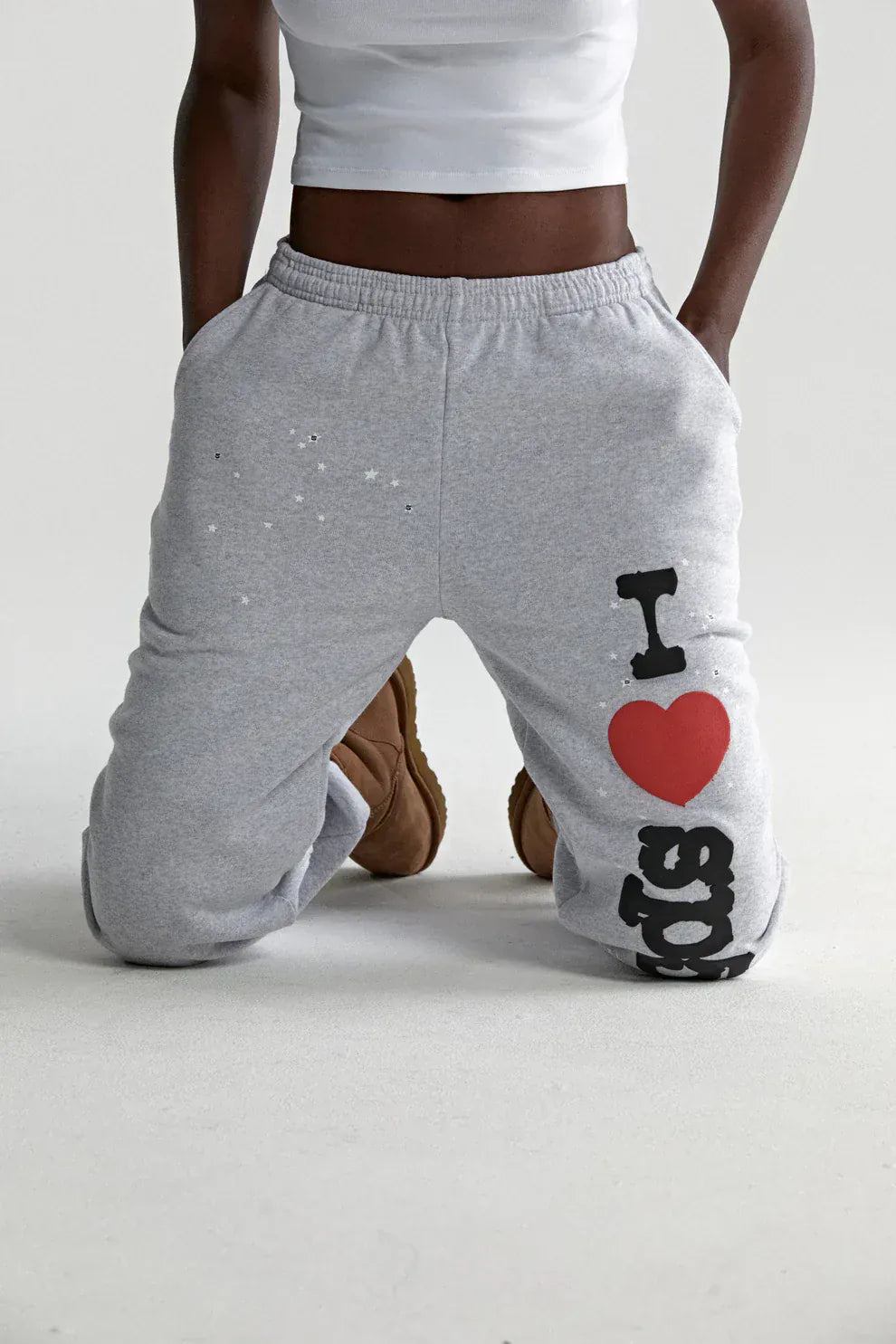 A person kneels on the floor wearing light grey 100% cotton Sp5der Sweatpants with an “I Love Sp5der” graphic, a white crop top, and brown boots.