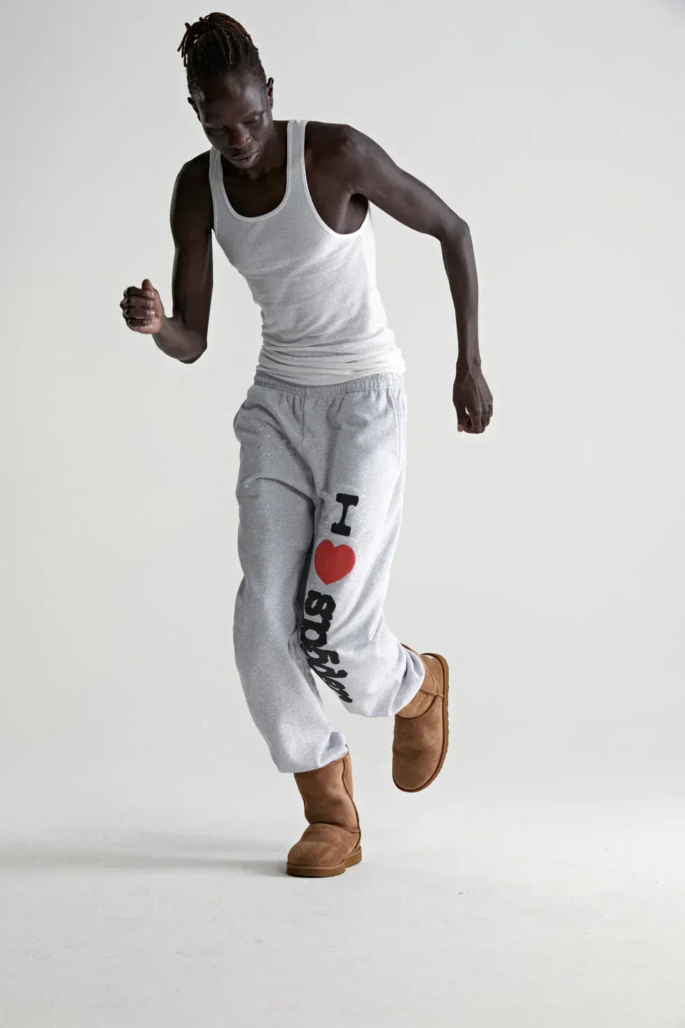 A person in a white tank top, tan boots, and 100% Cotton Sp5der Sweatpants for Men—light grey jogger pants featuring an “I Love Sp5der” graphic and relaxed fit—poses mid-step against a plain background.
