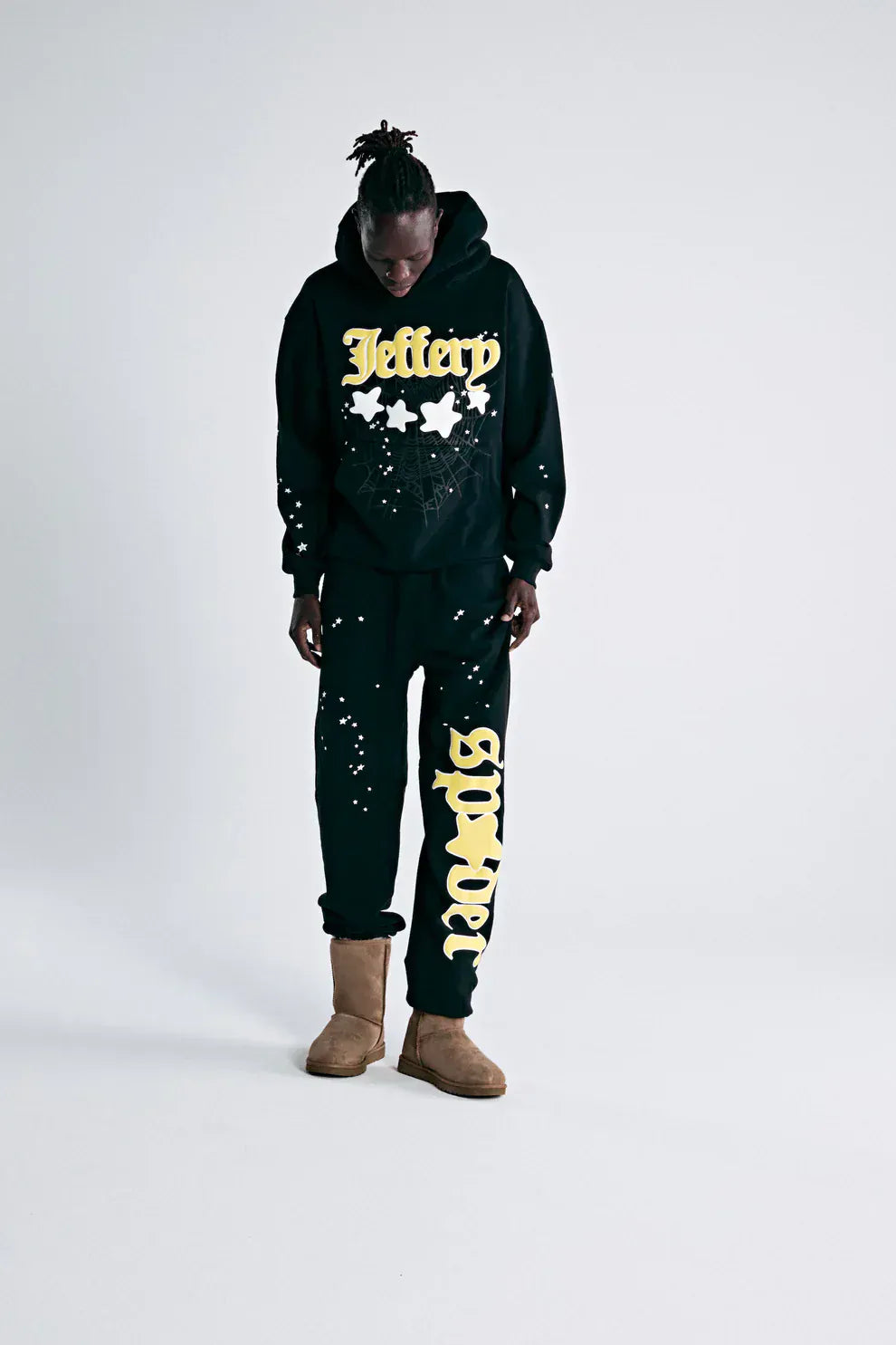 A person stands against a plain background wearing a black hoodie with "Jeffery" and stars, 100% Cotton Sp5der Sweatpants for Men—Black Streetwear Jogger Pants with Yellow Logo Graphic, Relaxed Fit, and light brown boots.