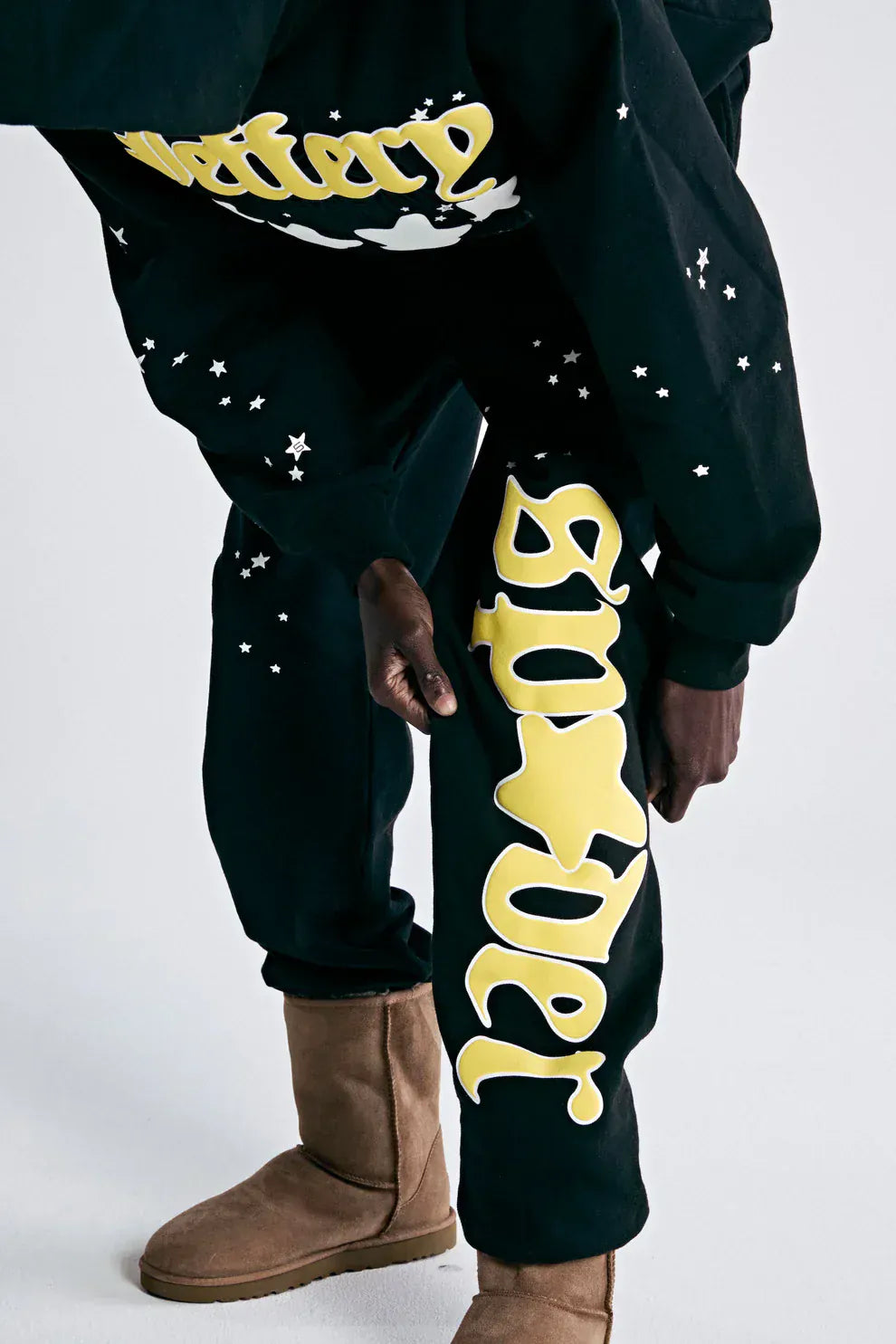 A person wears 100% Cotton Sp5der Sweatpants for Men—Black Streetwear Jogger Pants with Yellow Logo Graphic, relaxed fit—paired with tan boots, bending slightly to showcase these stylish joggers.