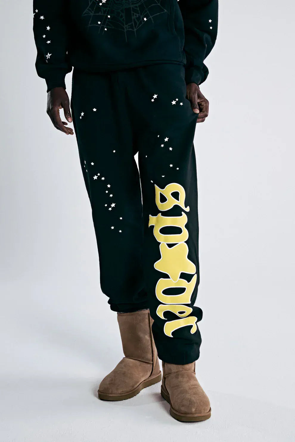 A person in 100% Cotton Sp5der Sweatpants for Men—black streetwear jogger pants with a yellow logo graphic and relaxed fit—paired with brown suede boots, creating a cool men's streetwear look.