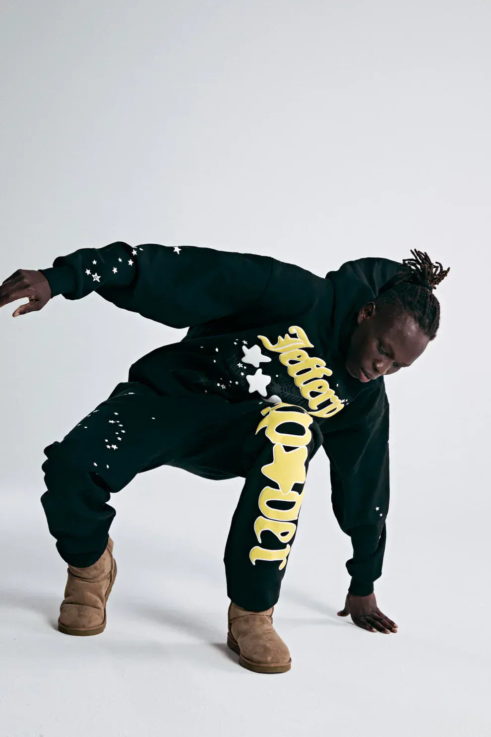 A person in a black hoodie and 100% Cotton Sp5der Sweatpants for Men—Black Streetwear Jogger Pants with Yellow Logo Graphic, Relaxed Fit—crouches in tan boots against a plain light background.