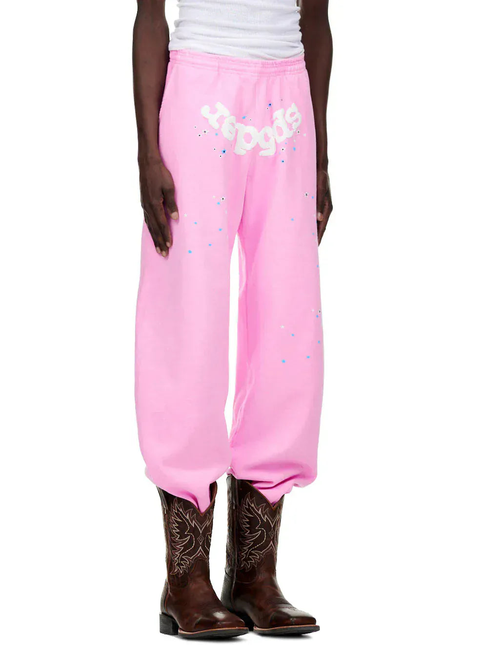 A person wearing Sp5der Sweatpants – Unisex Light Pink Cotton Sweatpants with front logo and star print, paired with a white tank top and brown cowboy boots, stands against a plain background.