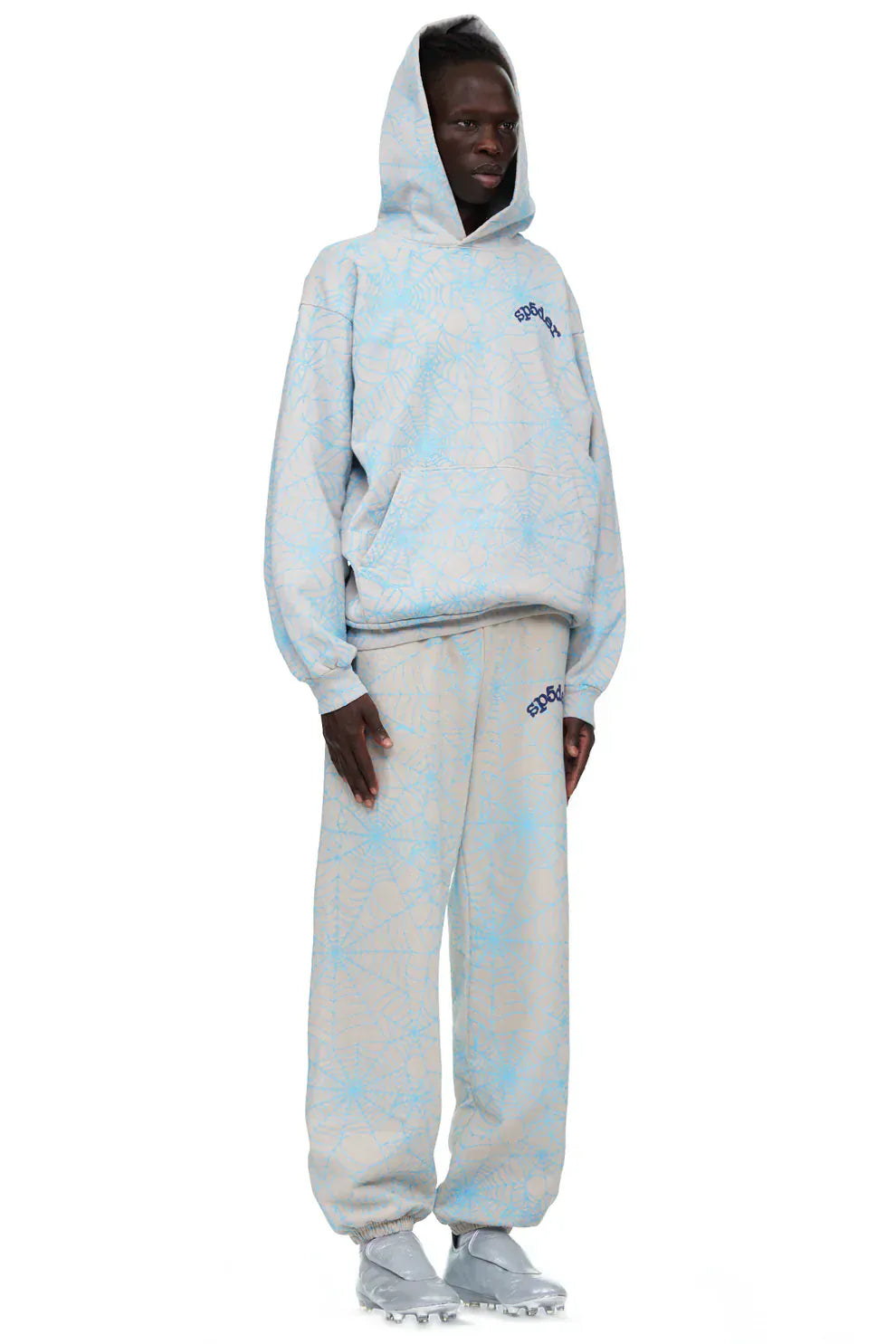 A person wearing a light blue hoodie, 100% Cotton Sp5der Sweatpants for Men in light grey with an all-over spider web print and relaxed fit, gray sneakers, and the hood up stands against a plain background.