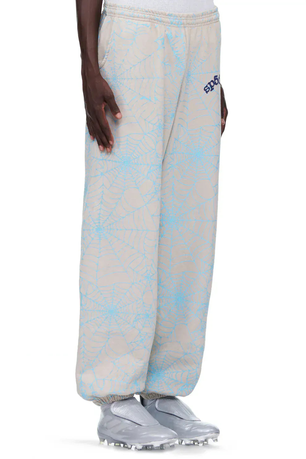A person wears 100% Cotton Sp5der Sweatpants for Men in light grey, featuring an all-over spider web print and relaxed fit, along with silver athletic shoes, standing against a plain background.