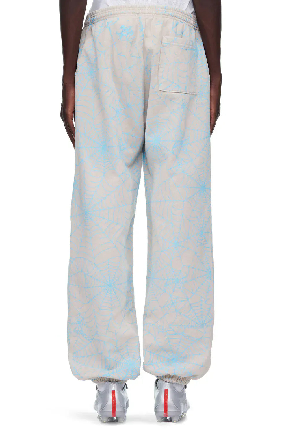 Model wears 100% Cotton Sp5der Sweatpants for Men in light grey with an all-over spider web print, relaxed fit, and elastic waistband and cuffs—styled from the back with white sneakers for a streetwear vibe.