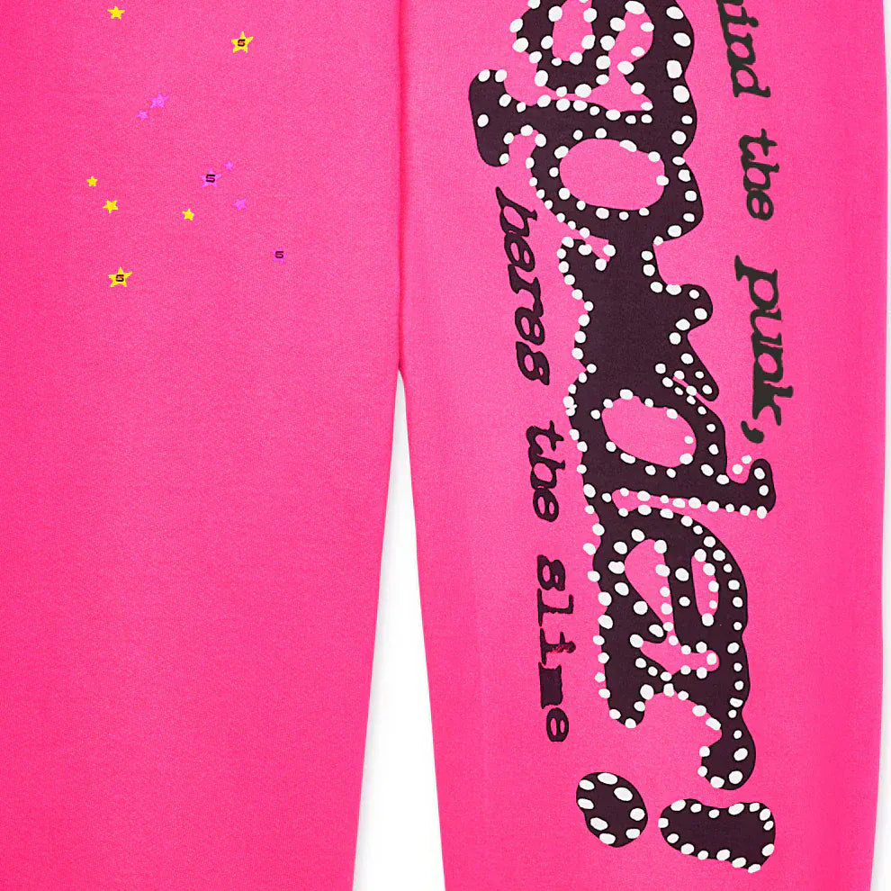 Close-up of Sp5der Sweatpants—unisex pink cotton wide leg style—featuring a graphic logo print, yellow and purple star decals, and bold black and white "spider! here’s the slime" text.