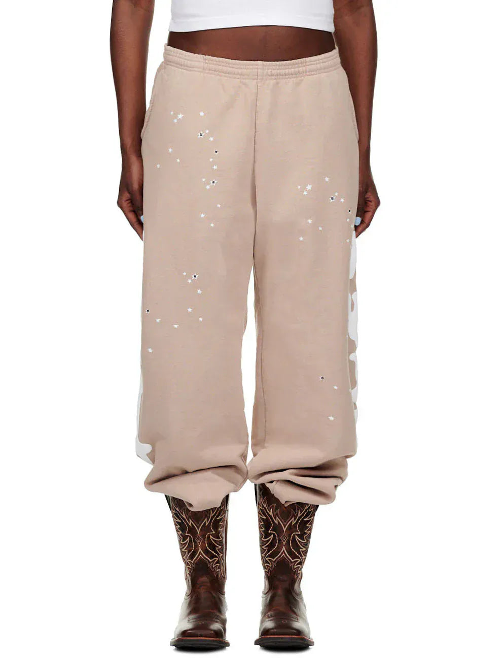 A person wears 100% Cotton Sp5der Sweatpants for Men—beige streetwear jogger pants with a side logo graphic and relaxed fit—paired with a white crop top and brown embroidered cowboy boots.